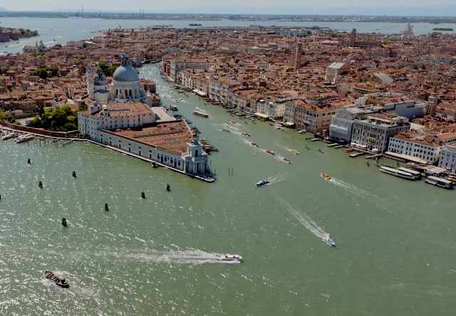 Aerial view of Venice, Italy, showing historic buildings, a domed church, and canals filled with boats on a sunny day.
