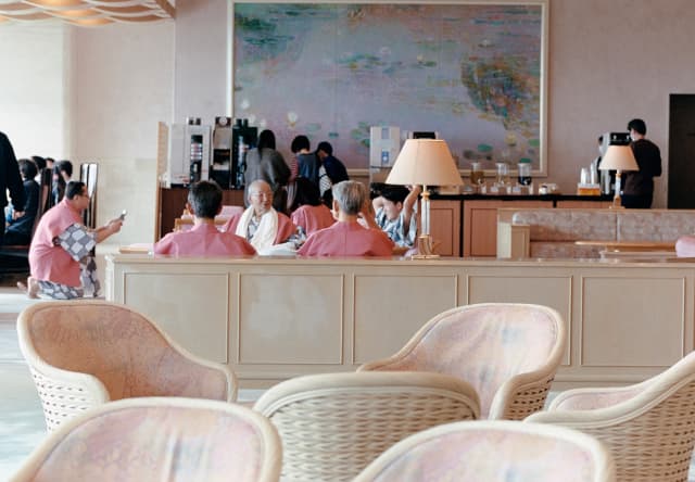 Five Japanese businessmen in pink tunics over grey-and-white tops relax at a table in a hotel, photographed by Cecy Young.