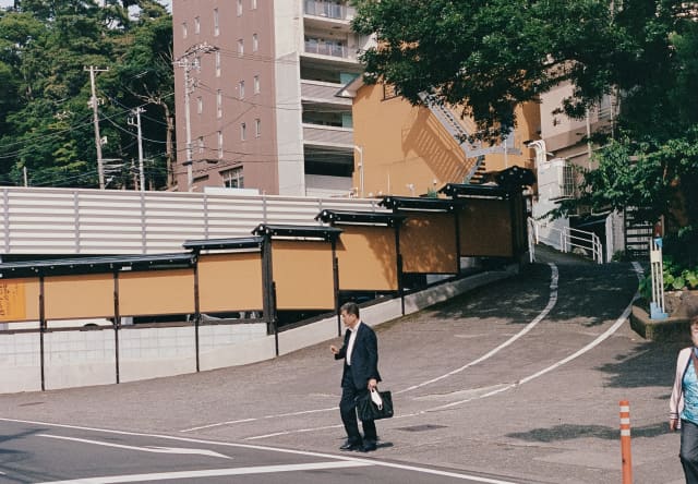 A businessman in a dark suit with a briefcase walks from blocks of apartments towards a wide road with crossing markings.