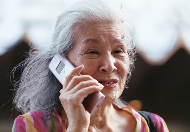An older Japanese woman with grey hair, minimal make-up and a pink-patterned top chats on a mobile phone, seen in close-up.