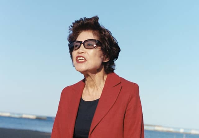 A Japanese woman with short black hair, wearing dark glasses and clothes with a red jacket, stands on a wide sunny beach.