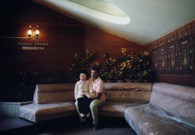 An Indian ventriloquist in a pink shirt and sage trousers sits on a large, curved banquette with his dummy on his knee.