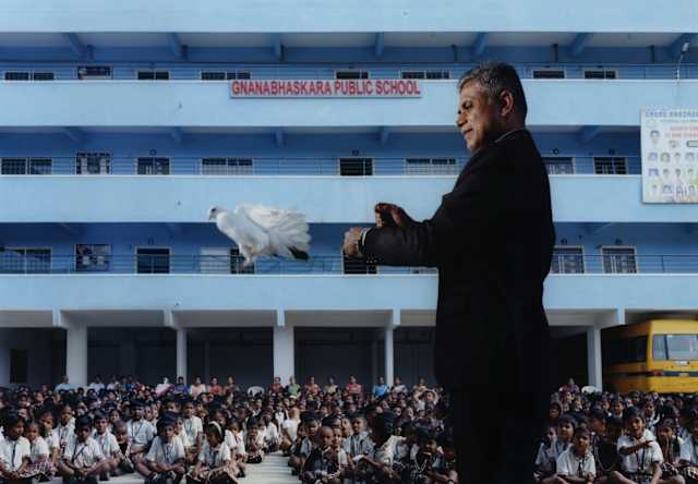 A magician in a black suit releases a white dove in front of a seated children in front of Gnanabhaskara Public School.