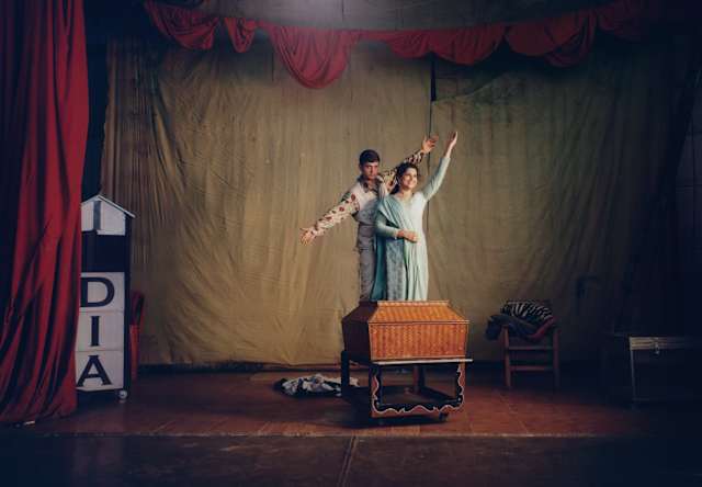 A woman and boy pose with a basket on a stage with a red curtain and broken 'India' sign at a magic convention in Bangalore.