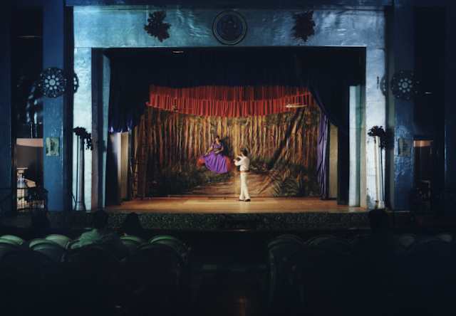 A magician in white levitates a woman in a purple on a red cushion on stage during The Great Mandrake show, seen from afar.