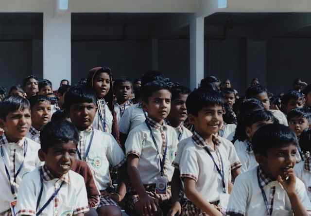 A group of school children with lanyards watch intensely as something happens off camera, in a close-up by Tara L. C. Sood.