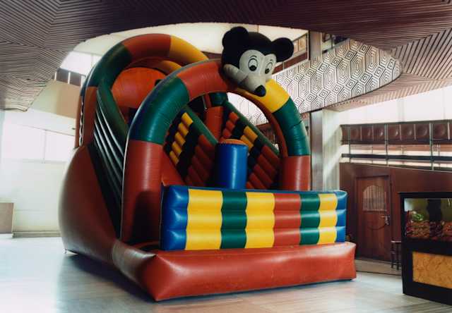 A red, blue, green and yellow inflatable slide with a Mickey Mouse-like figure fills a hall with an ornate carved mezzanine.