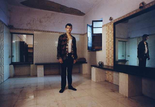 Magician Shankar Junior stands in an empty, tiled room in a maroon jacket and black trousers, reflected in a wall mirror.