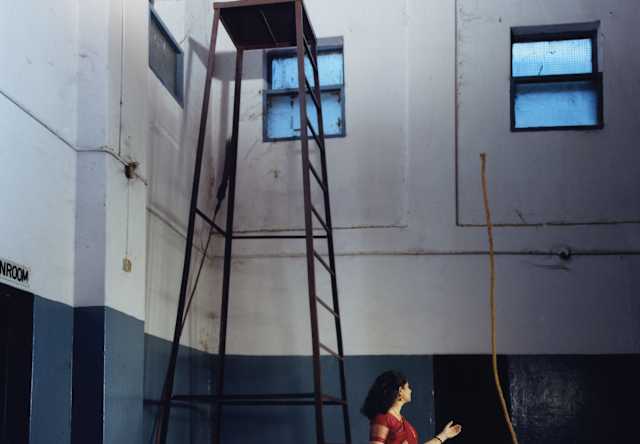 A woman with dark hair and a red dress levitates a rope so it stands high above her head by a tall platform in a large room.