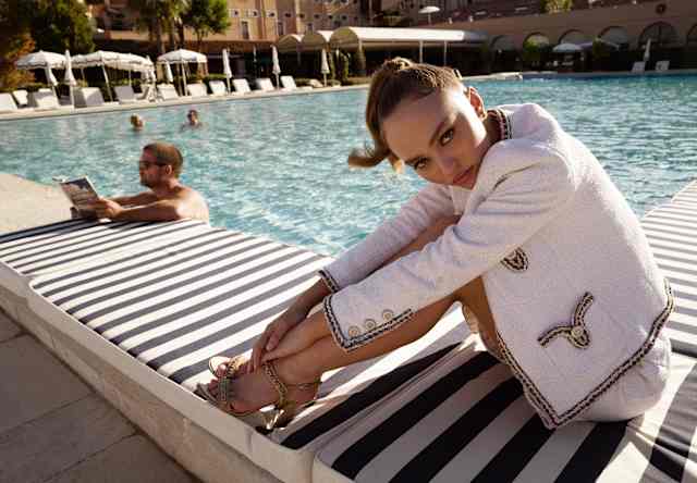 Lily-Rose Depp poses on the pool edge with hair tied back, wearing heels and a white Chanel jacket with black embroidering.