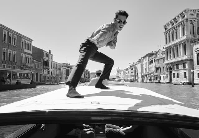 Rami Malek poses in socked feet and sunglasses on top of a water tax as it sails the Grand Canal, shot in black and white.