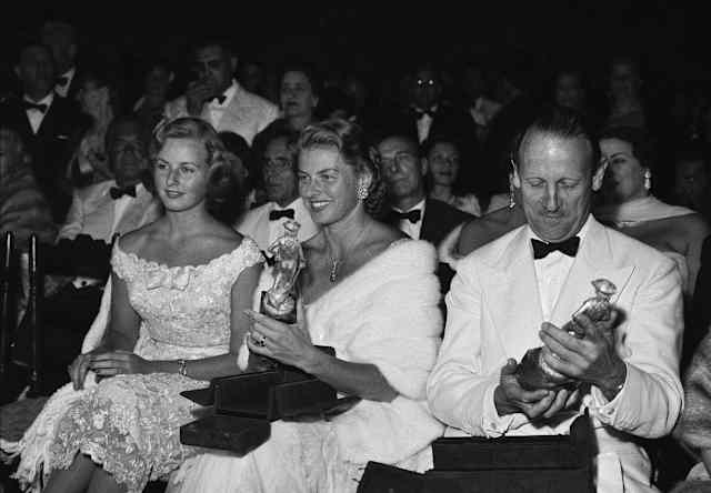 Ingrid Bergman, wearing white fur, and Sir Ashley Clarke hold their awards at the Taormina Film Festival, in black and white.
