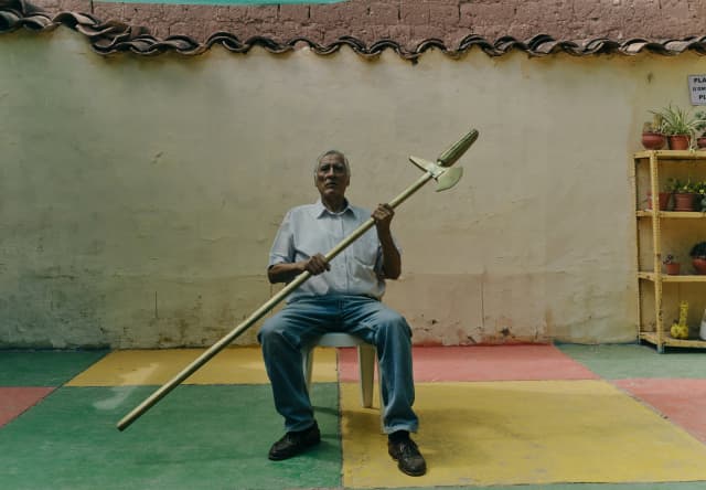 Alfredo Inca Roca, descendant of the Inca Viracocha, holds a golden spear in shirt and jeans, seated in a rustic courtyard.