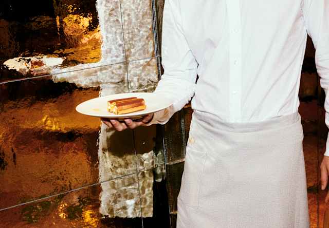 A waiter in a white shirt and beige apron holds a plate with a pudding, standing by a reflective, gold-toned wall.