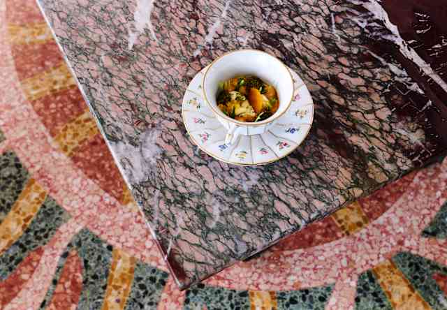 A small bowl of soup with herbs sits on a floral saucer atop a pink marble table, with a decorative mosaic floor visible below.