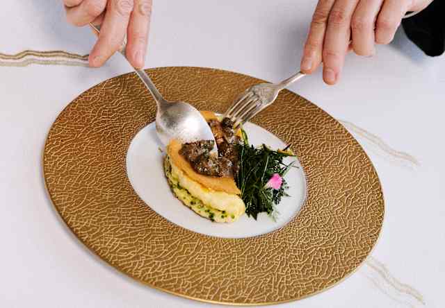 A person in formal attire cuts a small, gourmet dish on a gold-patterned plate, with greens on the side, atop a white tablecloth.