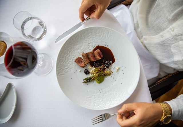 A diner holds a knife and fork by a plate of seared pork and asparagus at a table with red wine and water, seen from above.