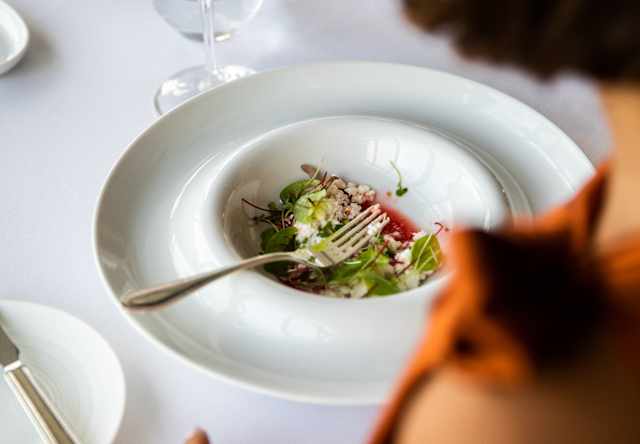 A fork rests on a moat-edged dish of beetroot with Requeijão cheese at a table with red wine, seen over a diner's shoulder.