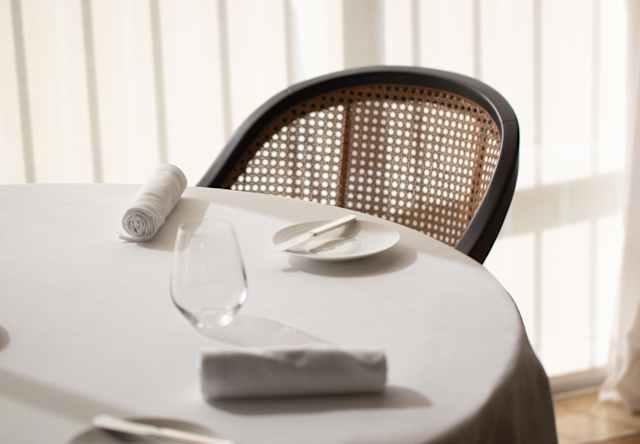 The curved back of a wood and wicker chair mirrors the sweep of a dining table edge in a detail, taken in sun-bleached light.