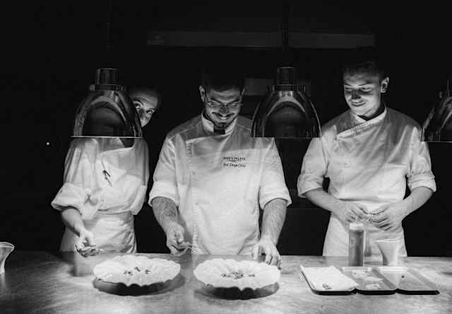 Behind a row of pendant heat lamps, Chef Costa and two chefs transfer garnish from trays to plates, seen in black and white.