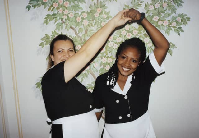 Two female staff members in black and white uniforms smile as as they twirl each other in a dance in front of a floral mural.