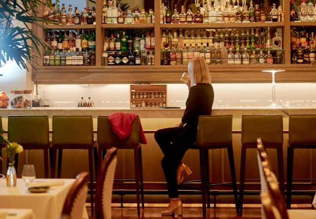 A woman sits alone at a bar with shelves full of bottles behind her. Elegant chandeliers hang from the ceiling, and warm lighting creates a cosy atmosphere in the stylish restaurant.