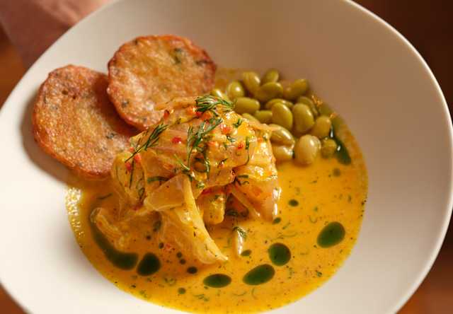 A white bowl holds a creamy yellow curry with pieces of chicken, garnished with green herbs and oil. Two toasted bread slices and a small serving of green beans are on the side. A person's hand holds the bowl.