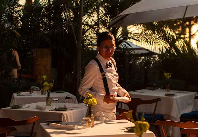 A waiter in a white shirt and apron stands beside elegantly laid tables with white tablecloths and flowers in vases at an outdoor restaurant during sunset.