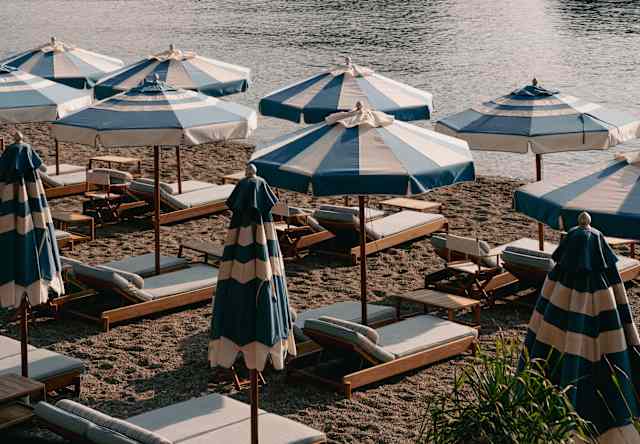 A shot of blue and white parasols on Mazzarò Bay beach at sundown, with views across still waters and boats to the headland.