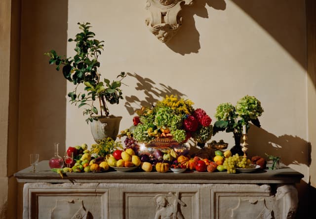 A stone altar-like table is adorned with a rainbow of produce including lemons, limes, pumpkins, grapes, cherries and flowers
