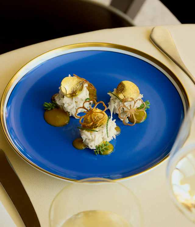An Oro dish of three mounds of white rice, topped with edible gold discs and served with sauce on a gilt-rimmed blue plate.