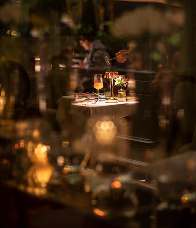 An orange cocktail sits in honeyed light on a dining table, seen through a haze of evening lights at Tragaluz restaurant.