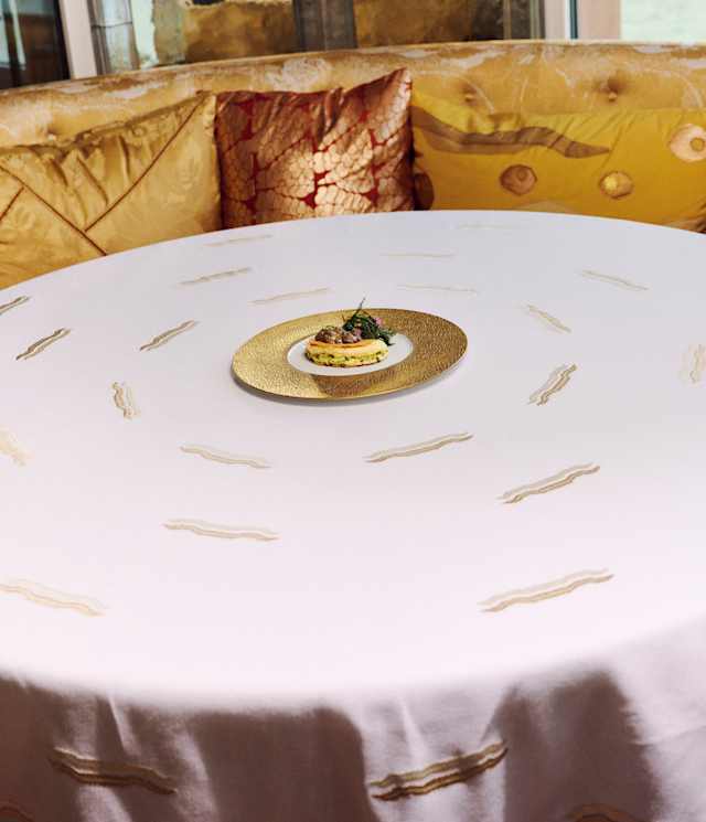 Smoked duck egg with a liver and morels served on a gilt-edged plate sits in the middle of a round table with gold-tone seating.