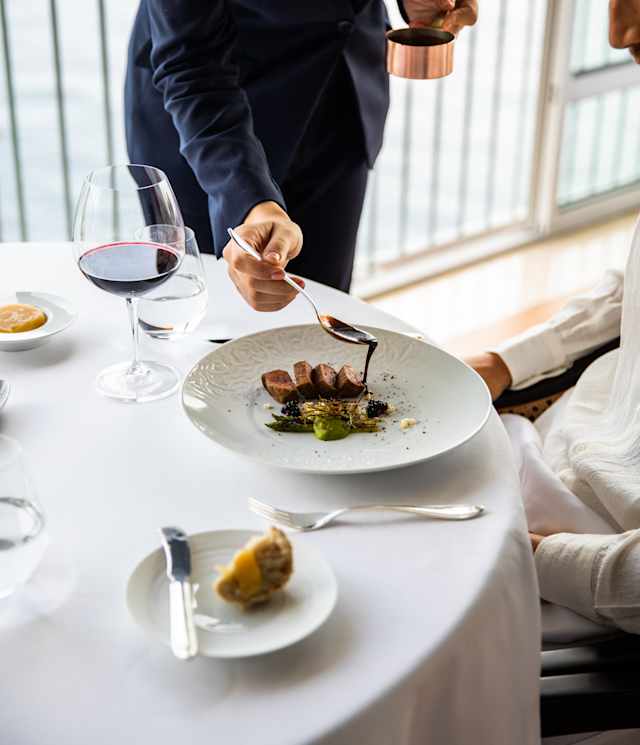 A diner watches a waitress spoon rich sauce over his plate of seared pork and asparagus at a table with a glass of red wine.