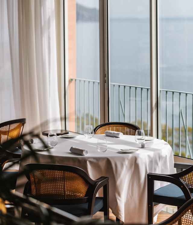 A window table with a white tablecloth, water glasses and napkins, and four cane chairs, claims a spectacular sea view.