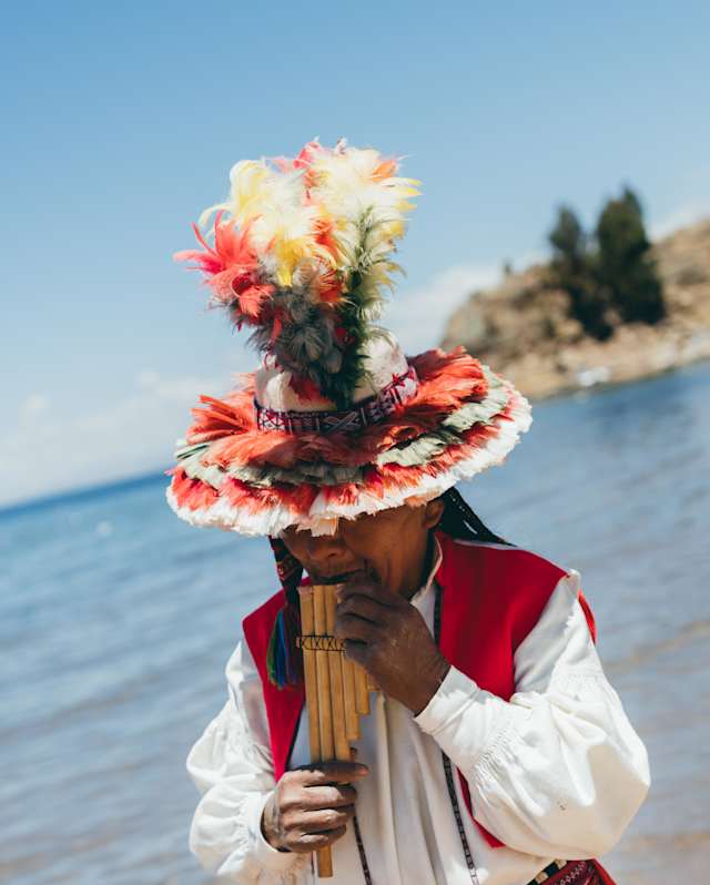 A person wearing traditional Andean clothing and a colourful feathered hat plays a pan flute by the water under a clear blue sky.