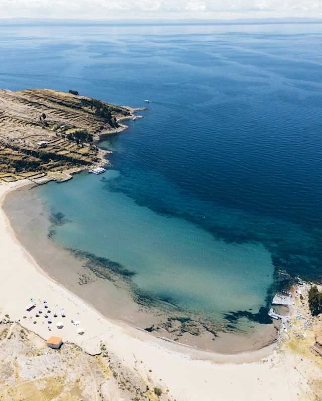 Aerial view of a sandy beach curving around a clear blue bay, with calm turquoise water, rocky coastline, and a vast open sea under a clear sky.
