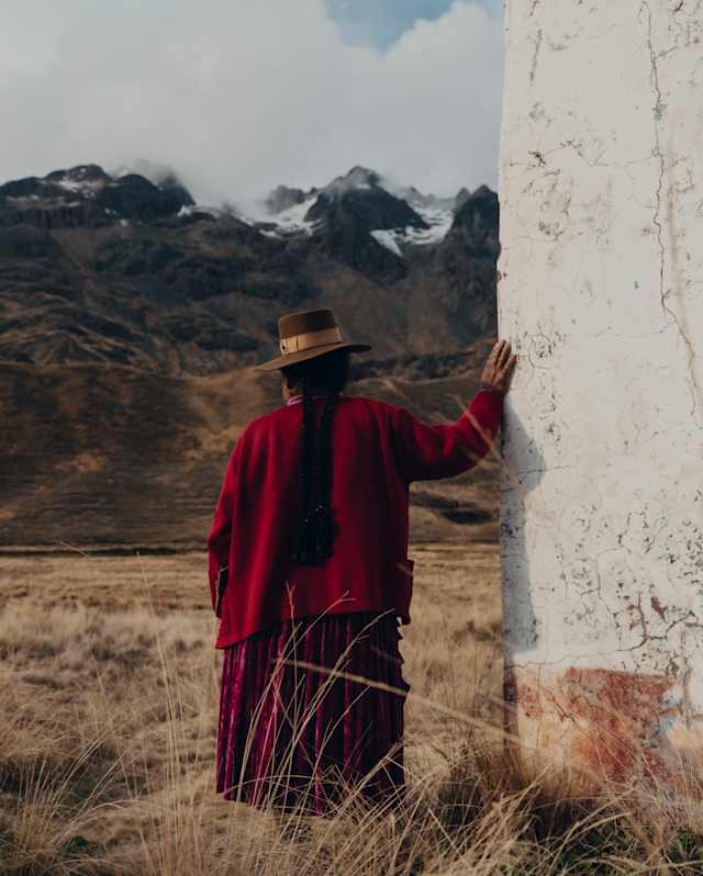A woman in a red clothing and a brown hat rests her hand on a white wall, with a snowy mountain backdrop, seen from behind.