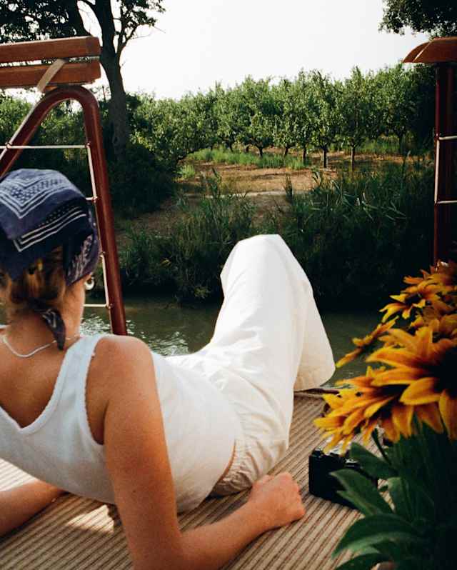 A woman in a white top and trousers with a blue headscarf lies back on the deck, gazing at the countryside, seen from behind.
