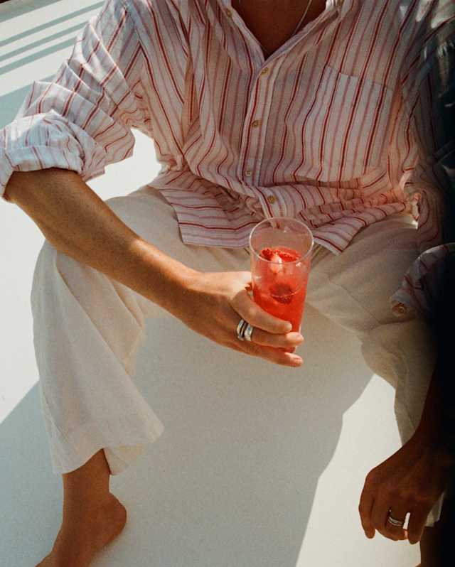 A guest, seen from the neck down in a red-striped shirt and white trousers, sits barefoot on the deck holding a pink drink.