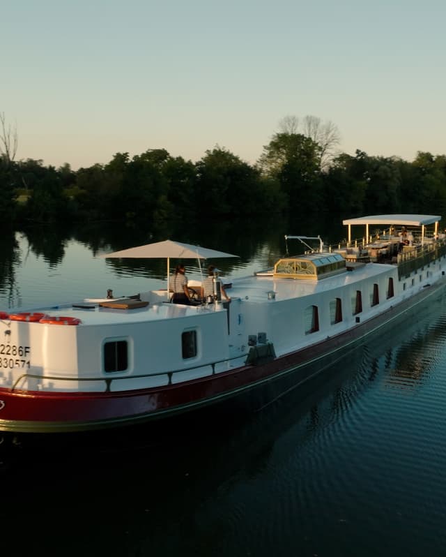 Soft sun brushes the canopy on the deck of Coquelicot as it glides along a wide, tree-lined waterway, seen from behind.