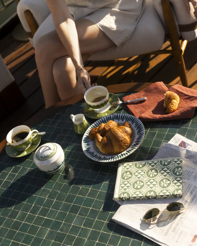 A woman sits at a low table with a book, paper, cups of tea and a bowl of croissants in an angled image of breakfast on deck.