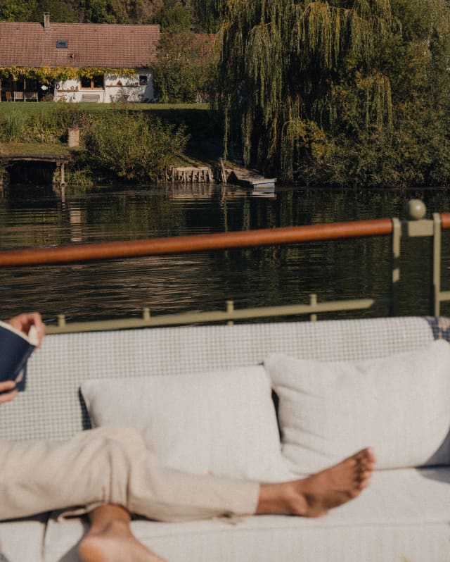 View over the legs of a male guest, reading on the deck sofa, to the riverbank, where willow branches bow into the waters.