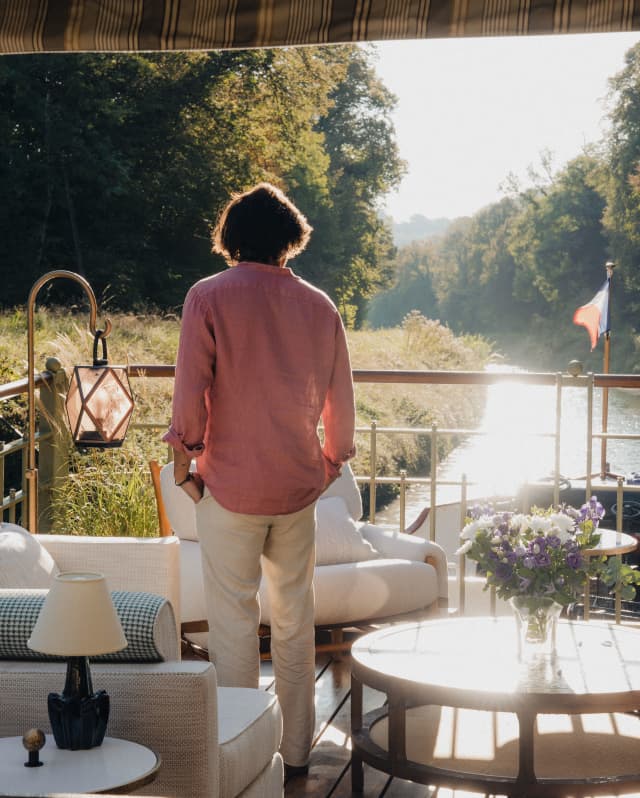 A man in a pink shirt and white trousers with dark hair watches the sun dance on the water from the deck, seen from behind.