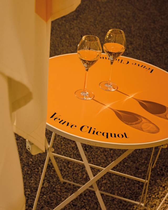 Two glasses of sparkling champagne cast their shadows on the vibrant orange top of a Veuve Cliequot-branded table.