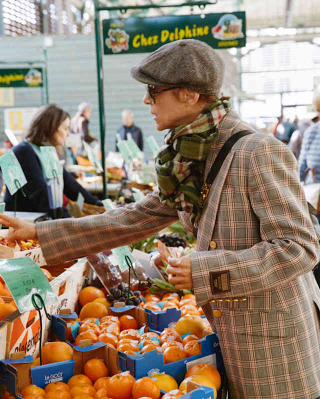 Chef Dominique Crenn, in checked jacket and flat cap, leans over orange crates as she selects produce at a market stall.