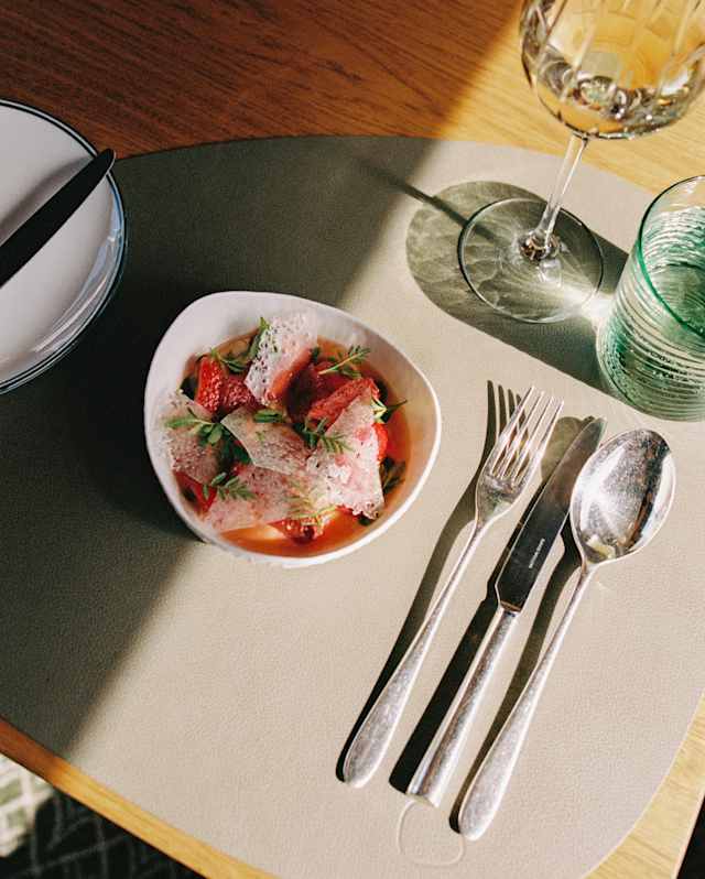 A small bowl of salad with greens and tomatoes sits on a placemat next to a fork, knife, and spoon, with a wine glass and a glass tumbler nearby on a wooden table. Sunlight partially illuminates the scene.