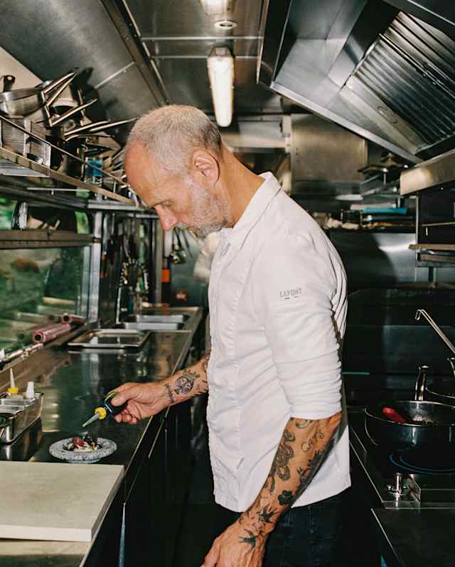 A chef with tattooed arms, wearing a white chef’s jacket, stands in a professional kitchen, holding a spoon and preparing food on a worktop, smiling or concentrating on his work.