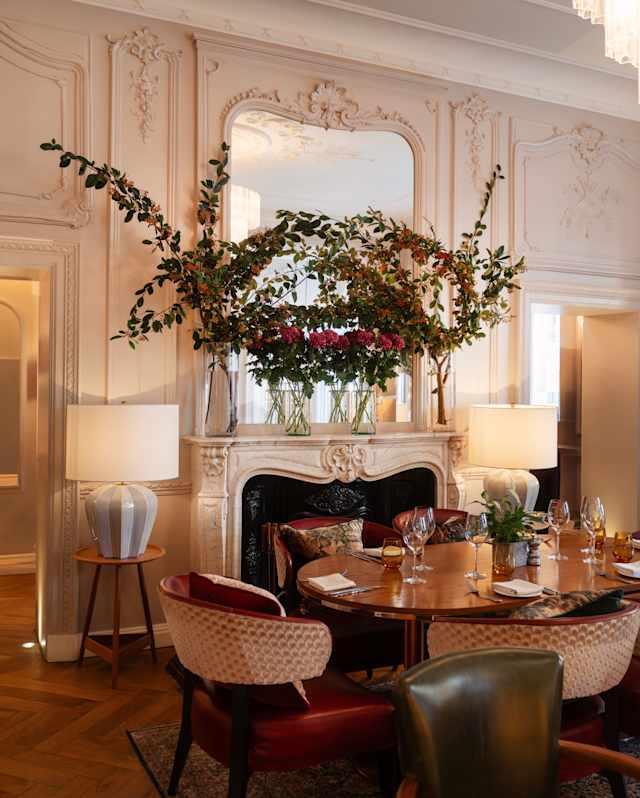 Two large glasses vases with flowers and foliage dominate a mirrored mantelpiece behind an oval dining table, laid for six.