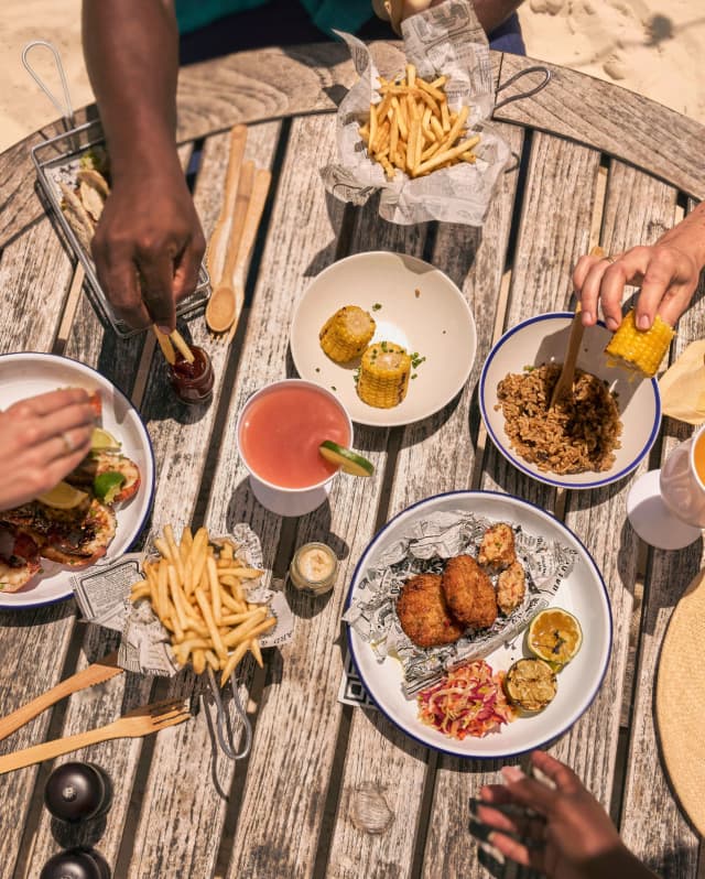 Diners' hands reach for grilled crayfish, corn, fries, and tasty plates at a Cap Shack table in the sand, seen from above.